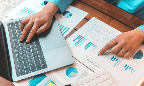 Person analyzing charts and graphs on printed documents while working on a laptop.