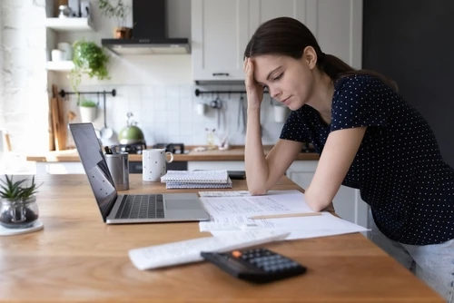 A young woman standing in her kitchen, leaning on the counter with her hand on her head, looking stressed. She is surrounded by bills, a calculator, and a laptop, as she ponders how much should I charge for rent.