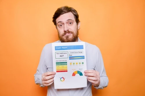 A man with a concerned expression holds up a credit report showing a low credit score of 427. The background is a solid orange color.