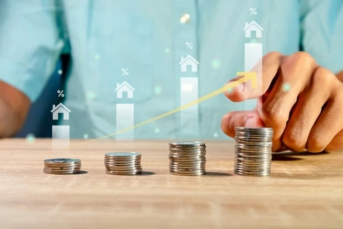 The image shows a person stacking coins in ascending order on a table, with graphic overlays of house icons and percentage symbols rising above the stacks. A yellow upward arrow connects the icons.