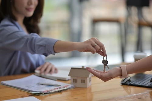 A person handing over house keys to another person across a desk with real estate documents and a house model, symbolizing a property transaction.