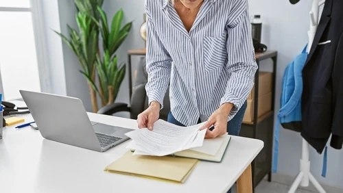 Woman shuffles paperwork on a desk next to an open laptop. 