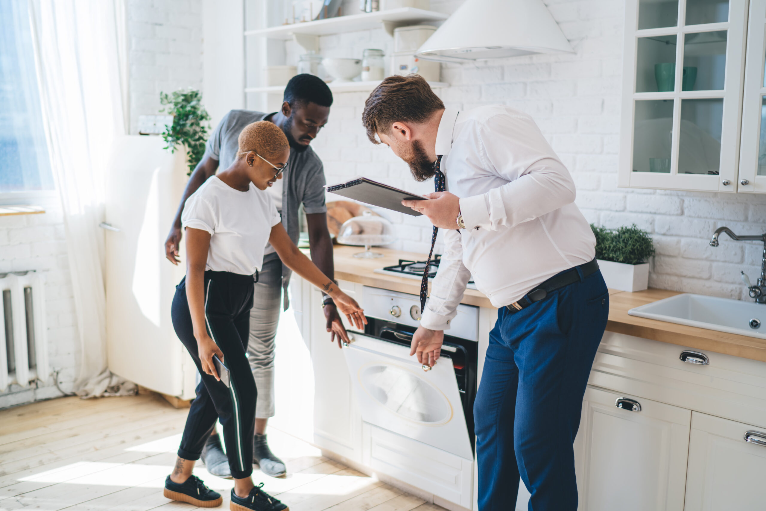 Professional man with clipboard inspecting oven together with attentive couple while standing at modern sunny apartment.
