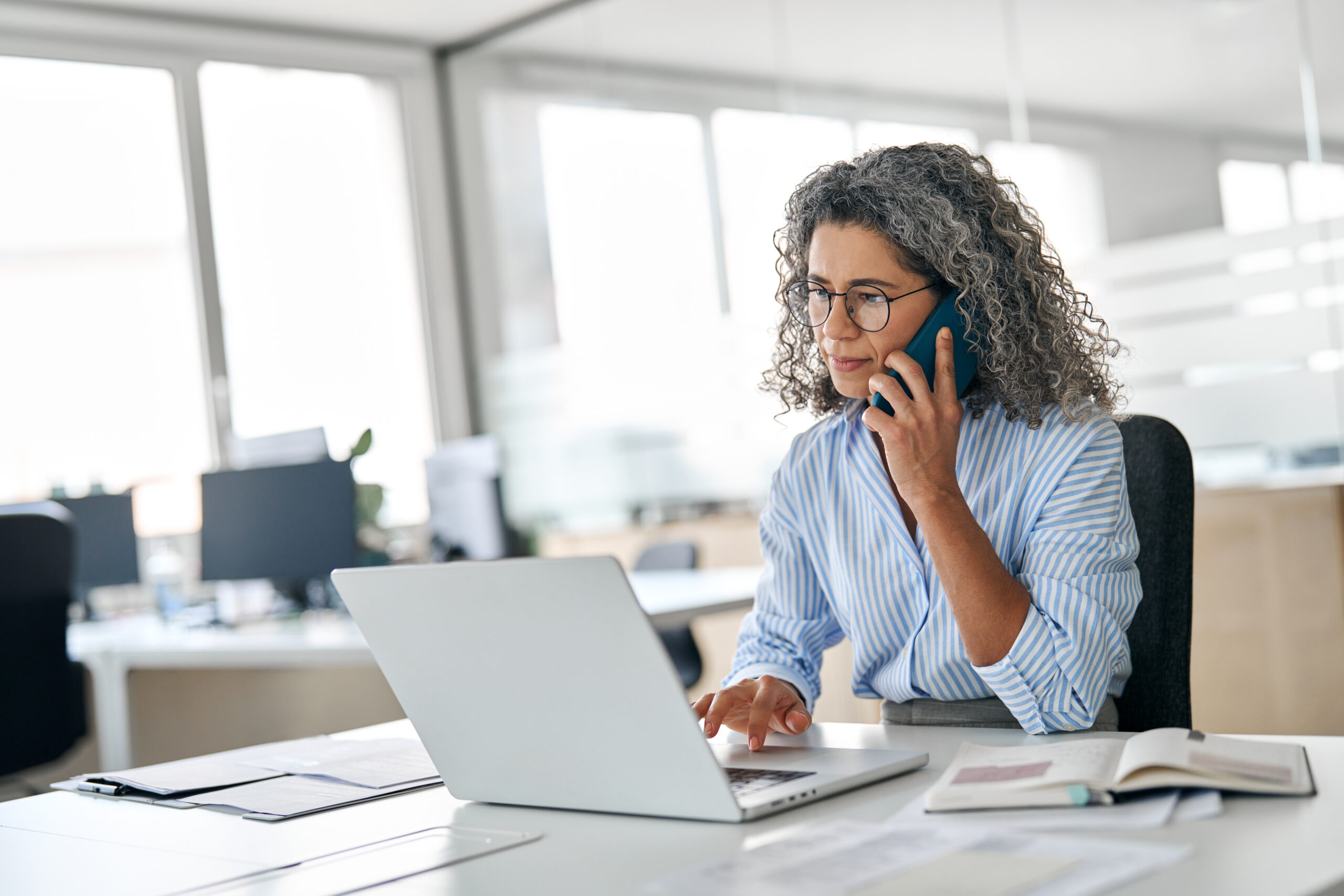 Busy middle aged business woman talking on the phone while sitting at work desk in office using laptop computer.