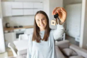 can you own a house and rent another​, cheerful female moved in new house, rent apartment, bought own real estate, the key with keyring in shape of little house